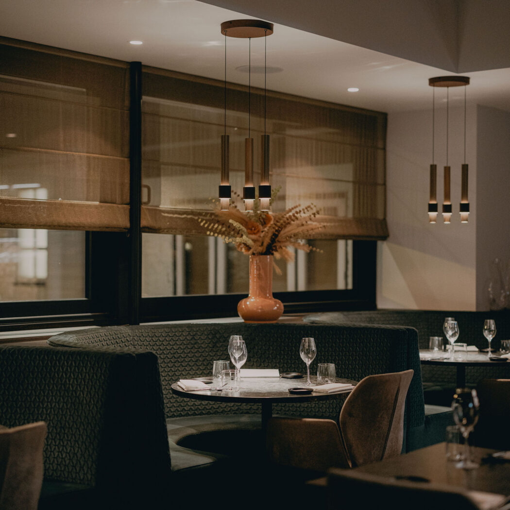 Sfeervol restaurantinterieur met donkergroene zitbanken, gedekte tafels, moderne hanglampen en oranje vaas.