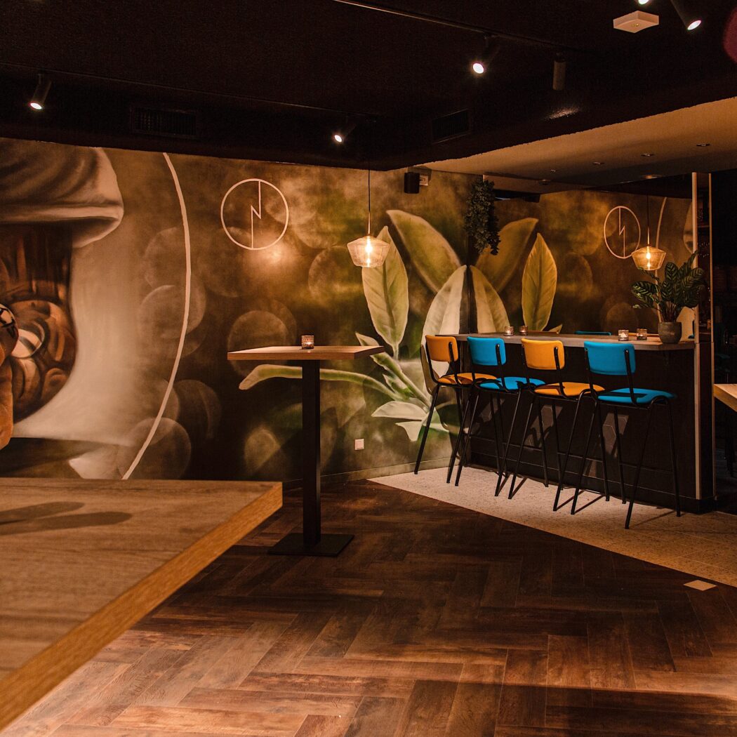 Sfeervol barinterieur met donkere visgraatvloer, botanische muurschildering, houten sta-tafels en felgekleurde barkrukken aan de bar.