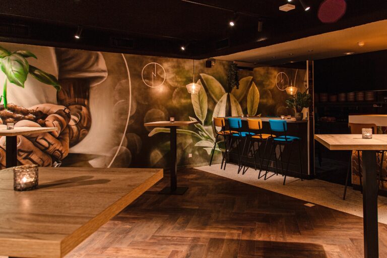 Sfeervol barinterieur met donkere visgraatvloer, botanische muurschildering, houten sta-tafels en felgekleurde barkrukken aan de bar.