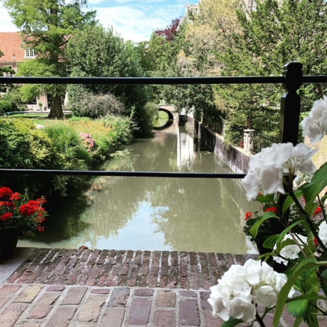 Zicht op een groene gracht met een boogbrug en omringende bomen, omkaderd door witte en rode bloemen.
