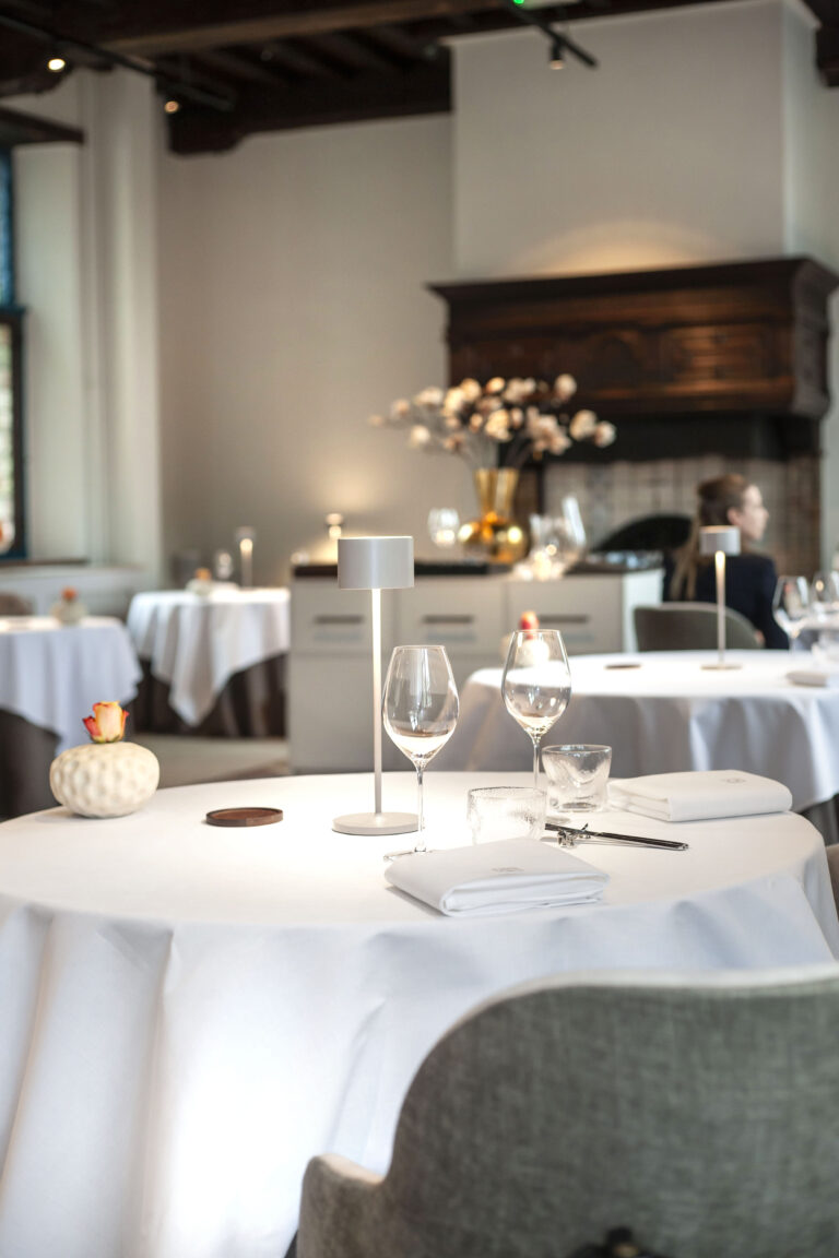 Gedekte tafel in een elegant restaurant met witte linnen, wijnglazen en een tafellampje, naast een oranje roos.