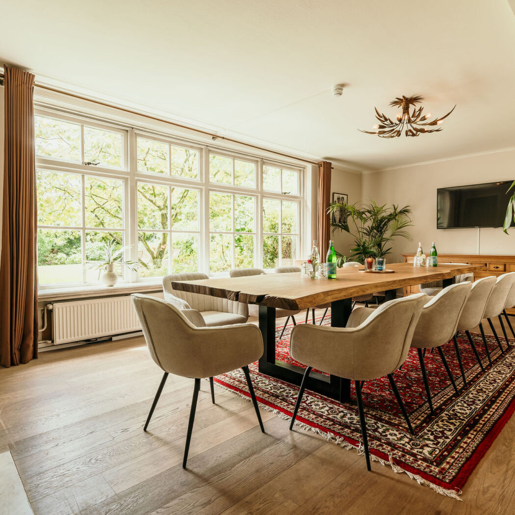 Lichte vergaderruimte met lange houten tafel, beige stoelen, groot raam met uitzicht op groen en een rood tapijt.