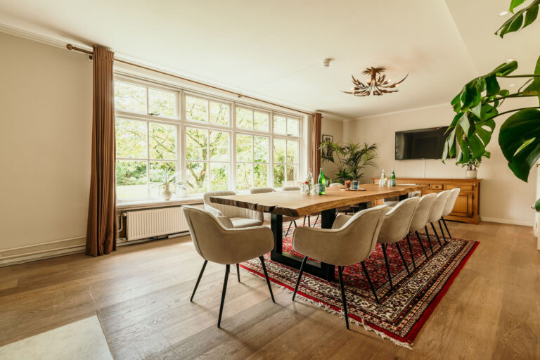 Lichte vergaderruimte met lange houten tafel, beige stoelen, groot raam met uitzicht op groen en een rood tapijt.