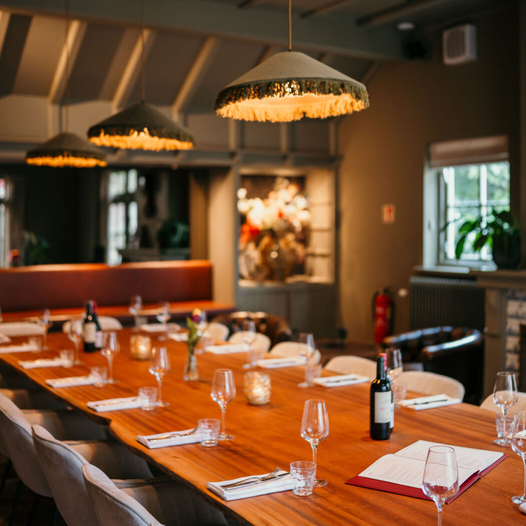 Lange houten eettafel met witte borden, glazen en wijn in een sfeervol restaurant met hanglampen.