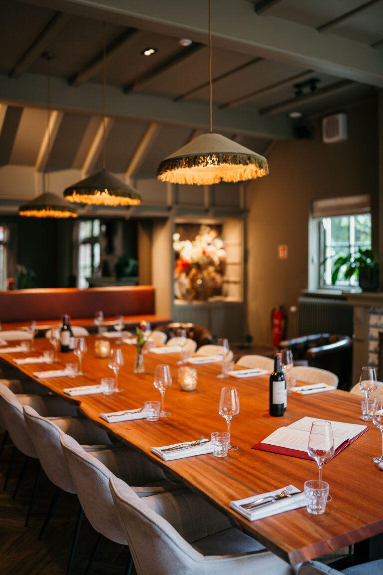 Lange houten eettafel met witte borden, glazen en wijn in een sfeervol restaurant met hanglampen.