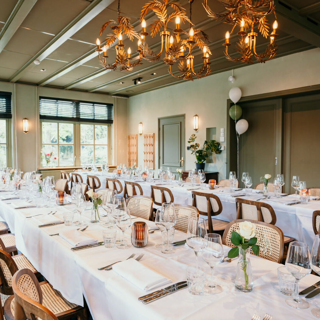 Lange tafels gedekt voor diner met rieten stoelen en gouden kroonluchters in een groene zaal.