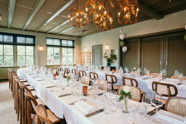 Lange tafels gedekt voor diner met rieten stoelen en gouden kroonluchters in een groene zaal.