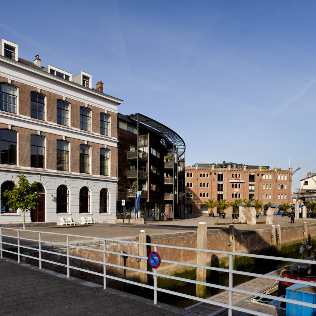Oud bakstenen gebouw aan een zonnige havenkade met boten, een moderne flat en een industriële kraan.