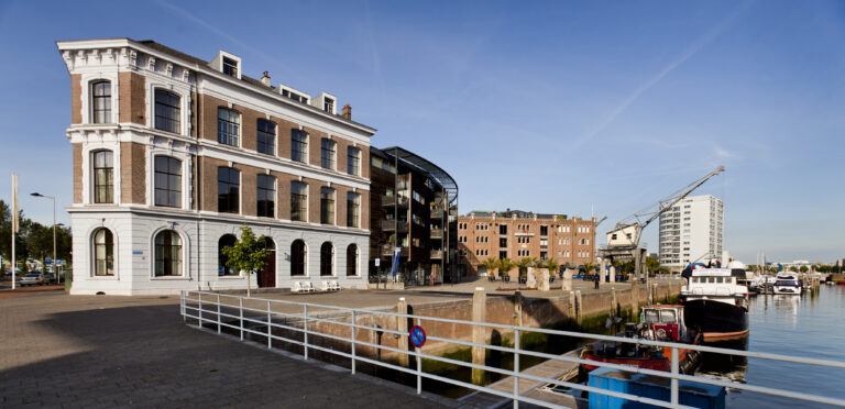 Oud bakstenen gebouw aan een zonnige havenkade met boten, een moderne flat en een industriële kraan.