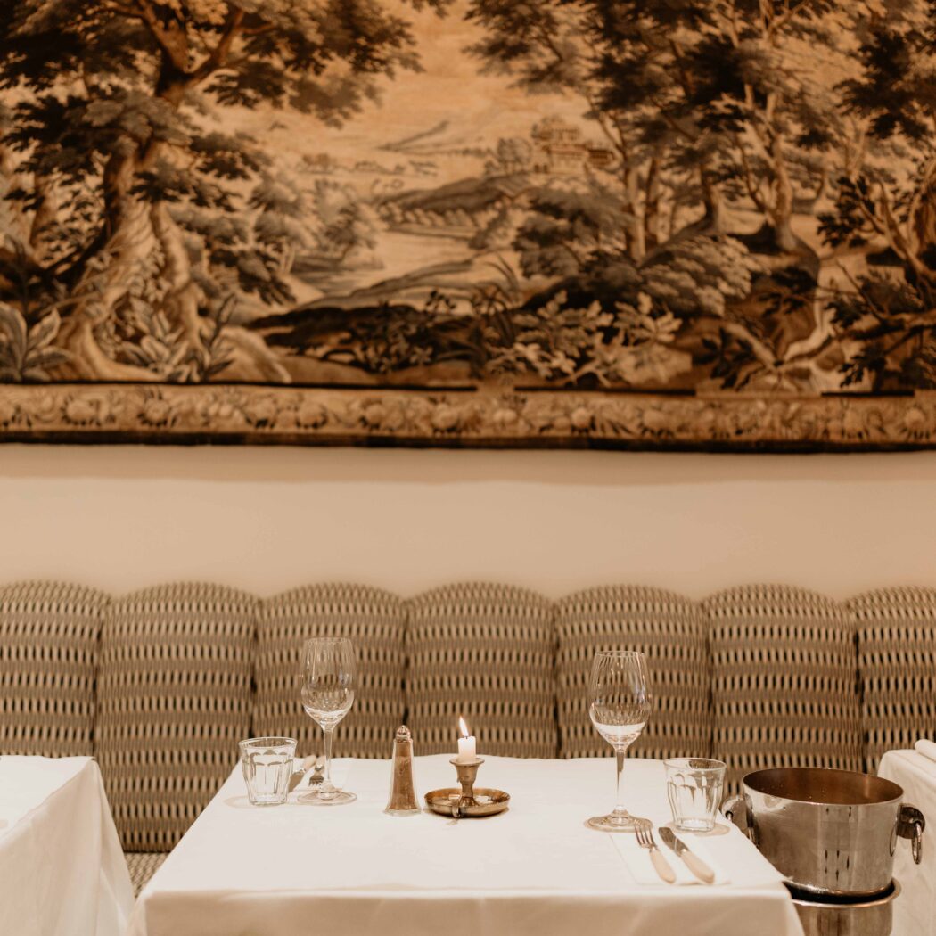 Elegant gedekte restauranttafel voor twee met brandende kaars, voor een patroonbank en groot landschapstapijt.