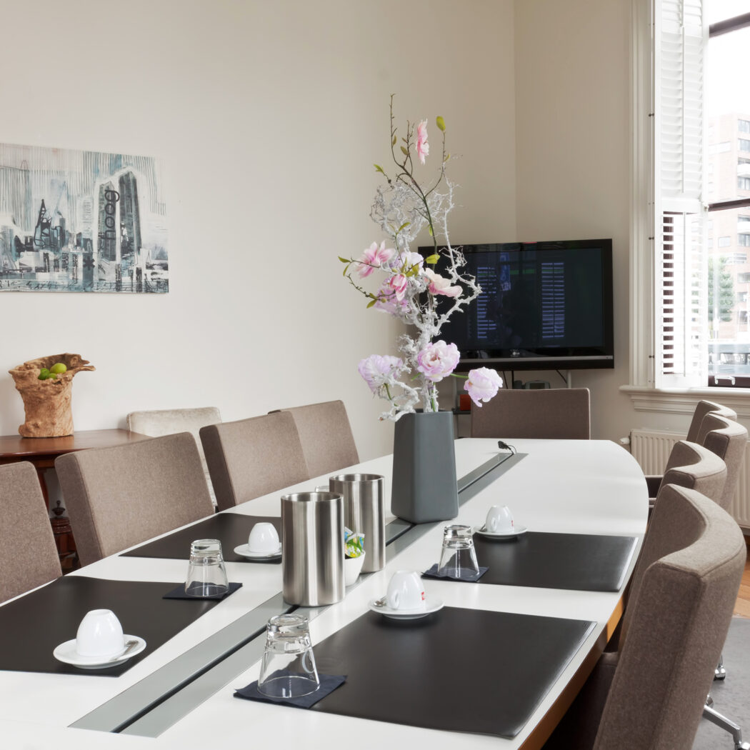 Stijlvolle vergaderruimte met lange witte tafel, beige stoelen, roze bloemstuk, tv en raam met witte shutters.
