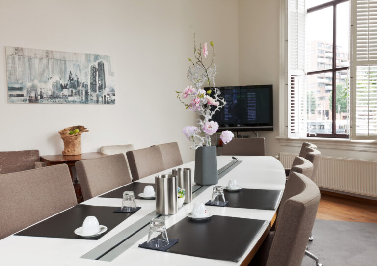 Stijlvolle vergaderruimte met lange witte tafel, beige stoelen, roze bloemstuk, tv en raam met witte shutters.