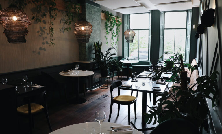 Sfeervol restaurantinterieur met donkergroene muren, hangplanten, geweven hanglampen en gedekte tafels.