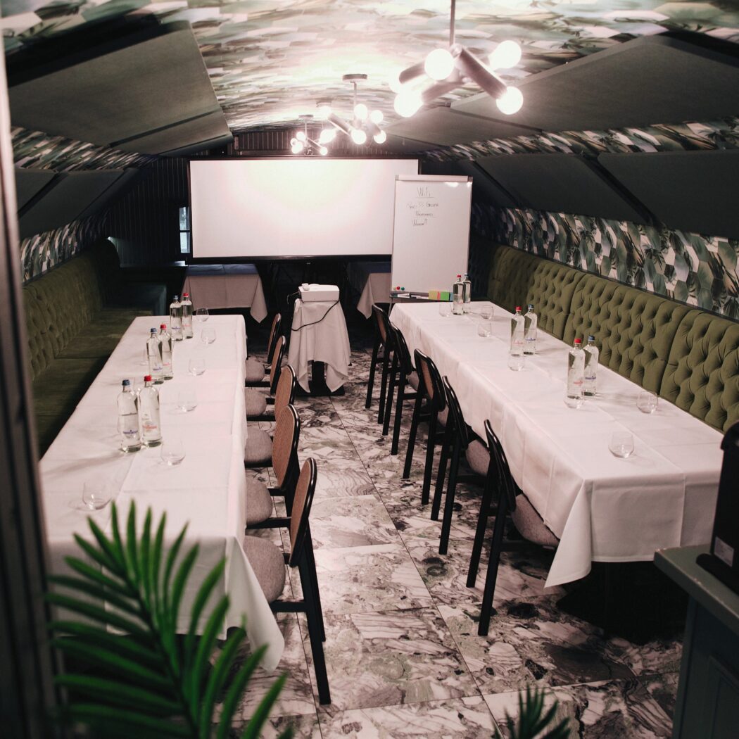 Stijlvolle vergaderzaal met lange tafels, groene banken, projectiescherm en bladmotief plafond.