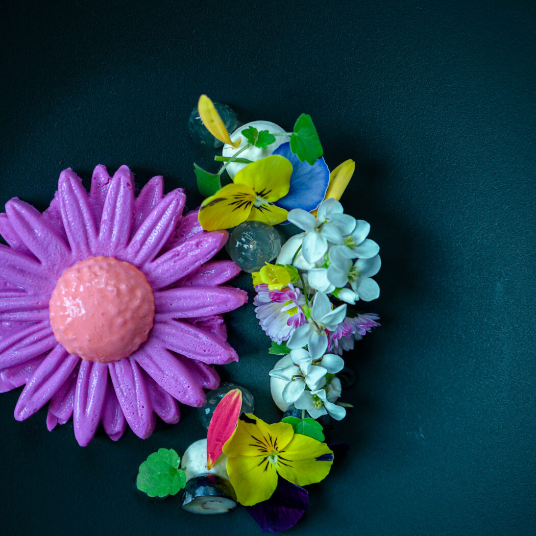 Bloemvormig paars dessert met perzikkleurig hart, omringd door kleurrijke eetbare bloemen op een donker bord.