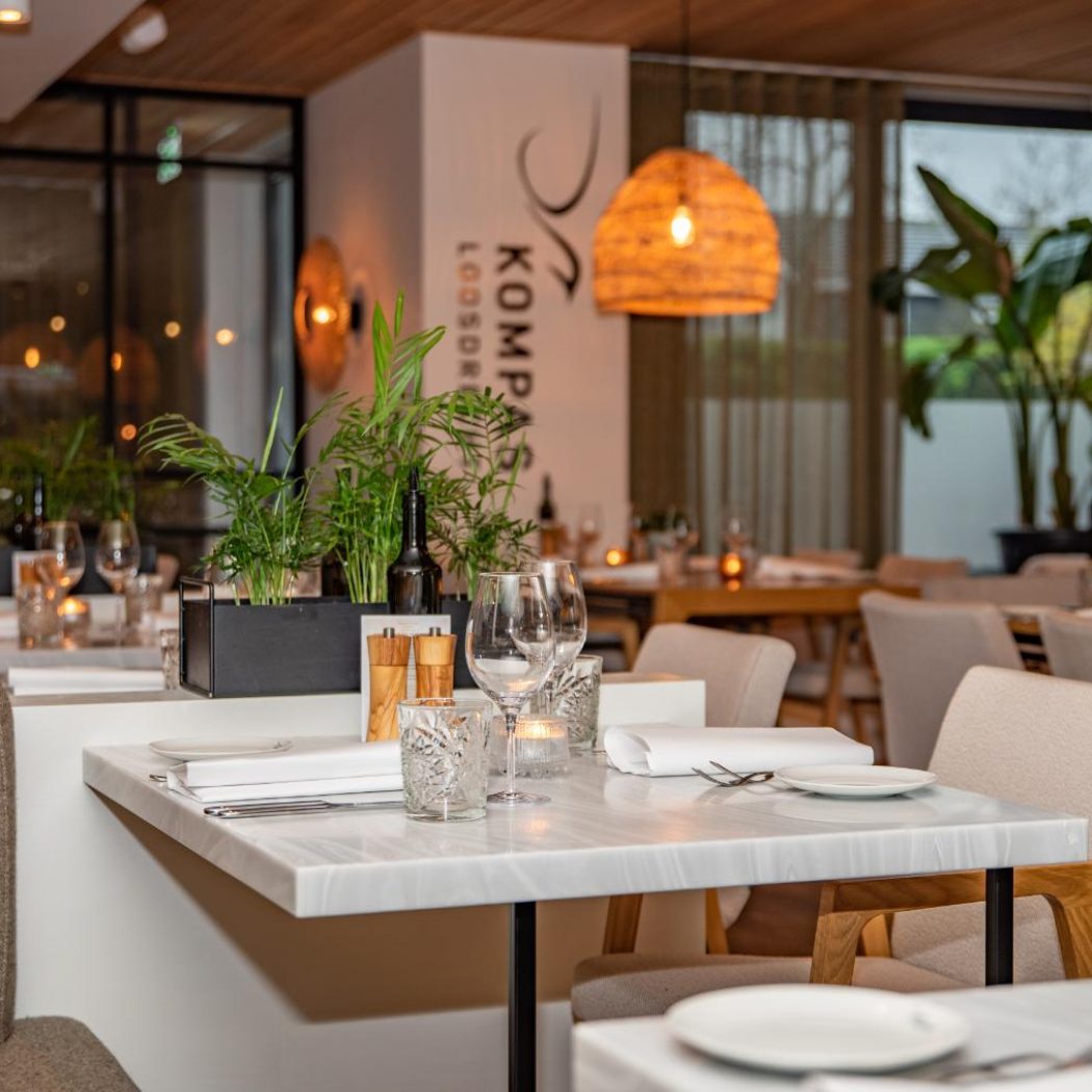 Modern restaurantinterieur met gedekte marmeren tafels, planten en het Kompas Loosdrecht logo op een witte zuil.