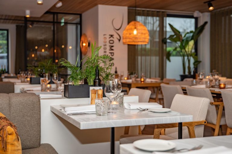 Modern restaurantinterieur met gedekte marmeren tafels, planten en het Kompas Loosdrecht logo op een witte zuil.