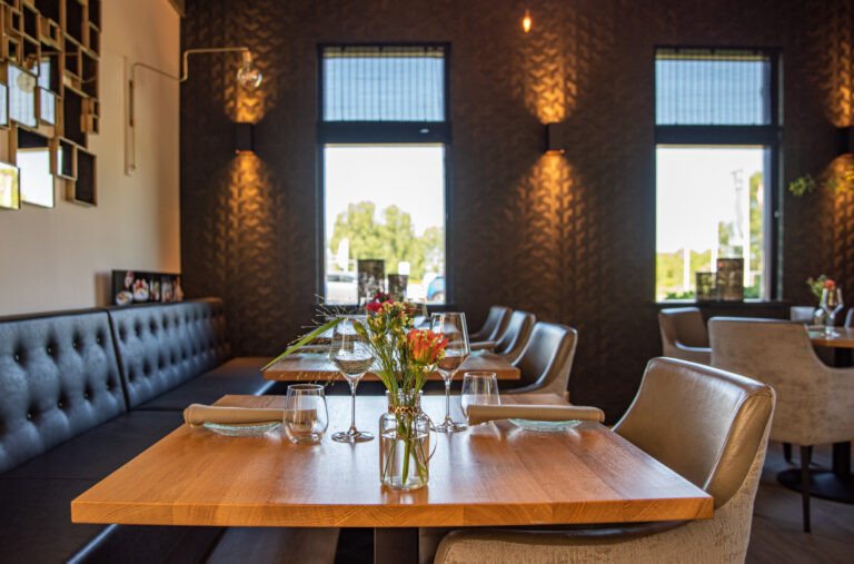 Sfeervol restaurant interieur met houten tafels, donkere banken, beige stoelen en bloemdecoratie bij de ramen.