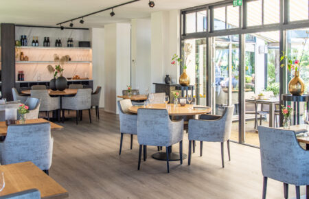 Lichte, moderne eetzaal met ronde houten tafels, grijze stoelen en grote ramen naar een buitenterras.