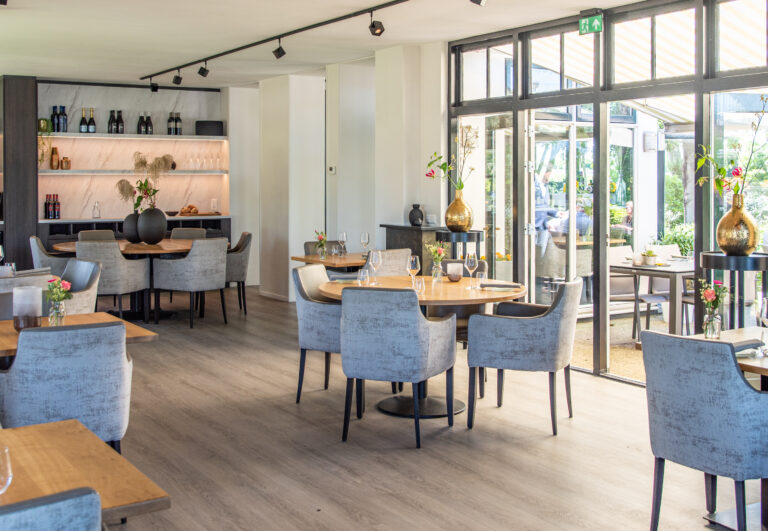 Lichte, moderne eetzaal met ronde houten tafels, grijze stoelen en grote ramen naar een buitenterras.