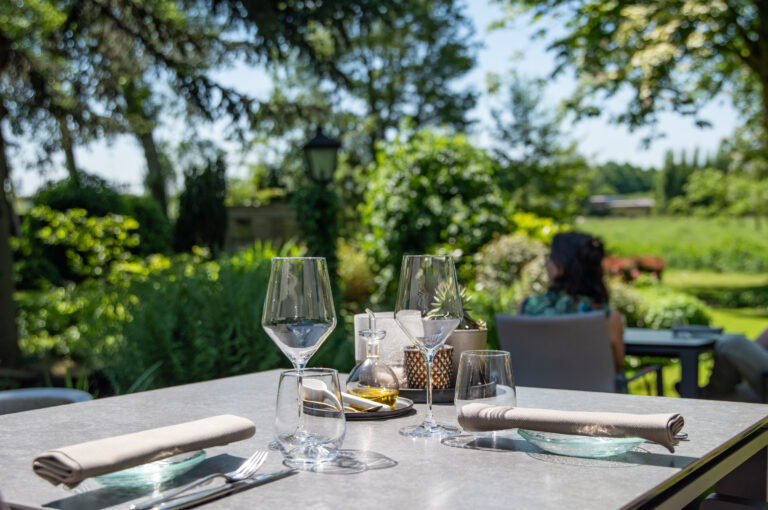 Buitentafel gedekt met glazen, bestek en servetten, met uitzicht op een weelderige tuin en een zittende gast.