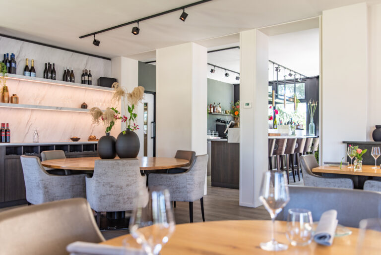 Licht restaurant interieur met ronde houten tafels, grijze stoelen, decoratieve vazen en verlichte wijnrekken.