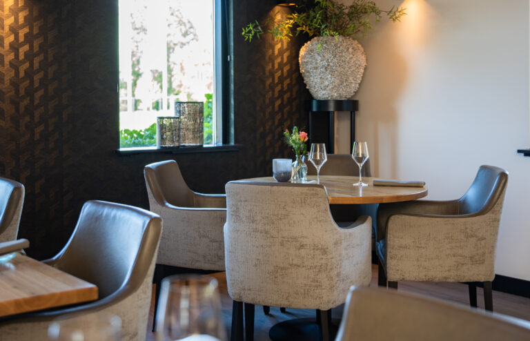 Ronde houten restauranttafel met vier comfortabele stoelen, gedekt met wijnglazen en bloemen.