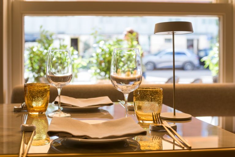 Gedekte restauranttafel met servies, twee gouden glazen, een tafellamp en raamzicht op een groene straat.
