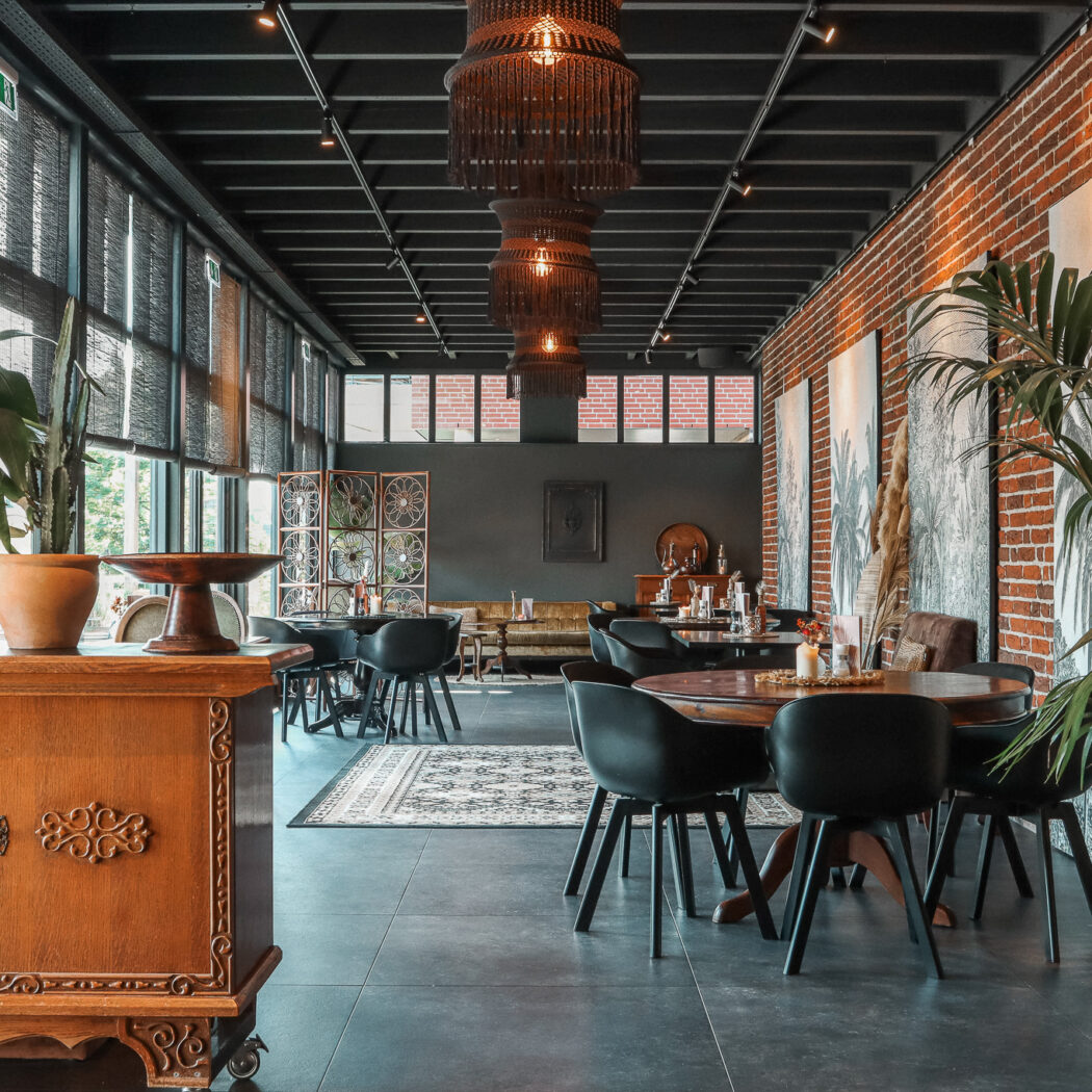 Sfeervol restaurant met donkere houten tafels, zwarte stoelen, bakstenen muur en veel planten bij de ramen.