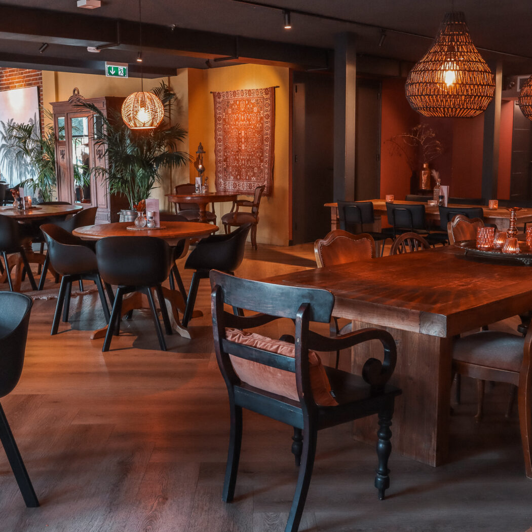 Sfeervol restaurantinterieur met houten tafels, diverse stoelen, rieten hanglampen en planten in warm licht.
