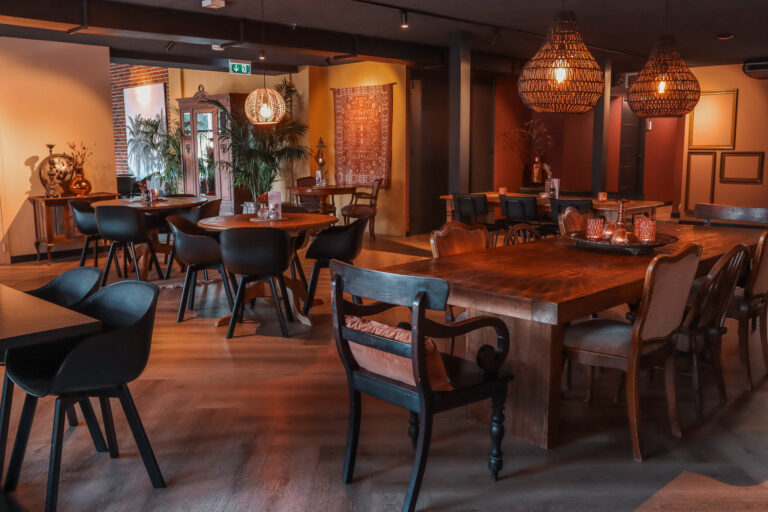 Sfeervol restaurantinterieur met houten tafels, diverse stoelen, rieten hanglampen en planten in warm licht.