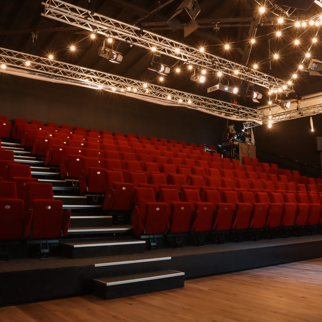 Lege theaterzaal met oplopende rijen rode stoelen, houten podium en sfeervolle podiumverlichting.