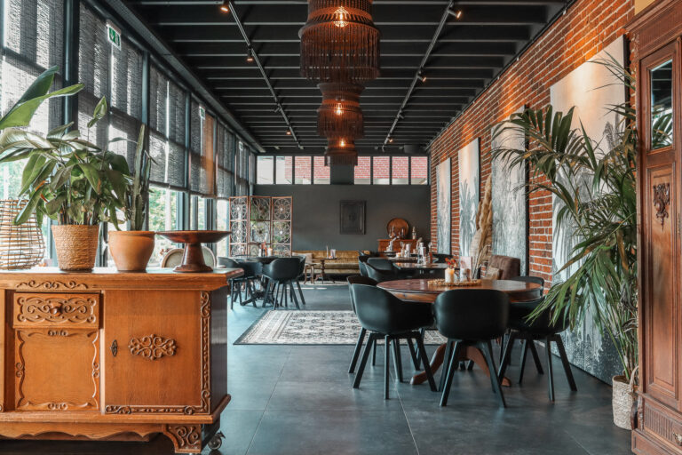 Sfeervol restaurant met bakstenen muur, donkere houten tafels, moderne stoelen en planten bij de hoge ramen.