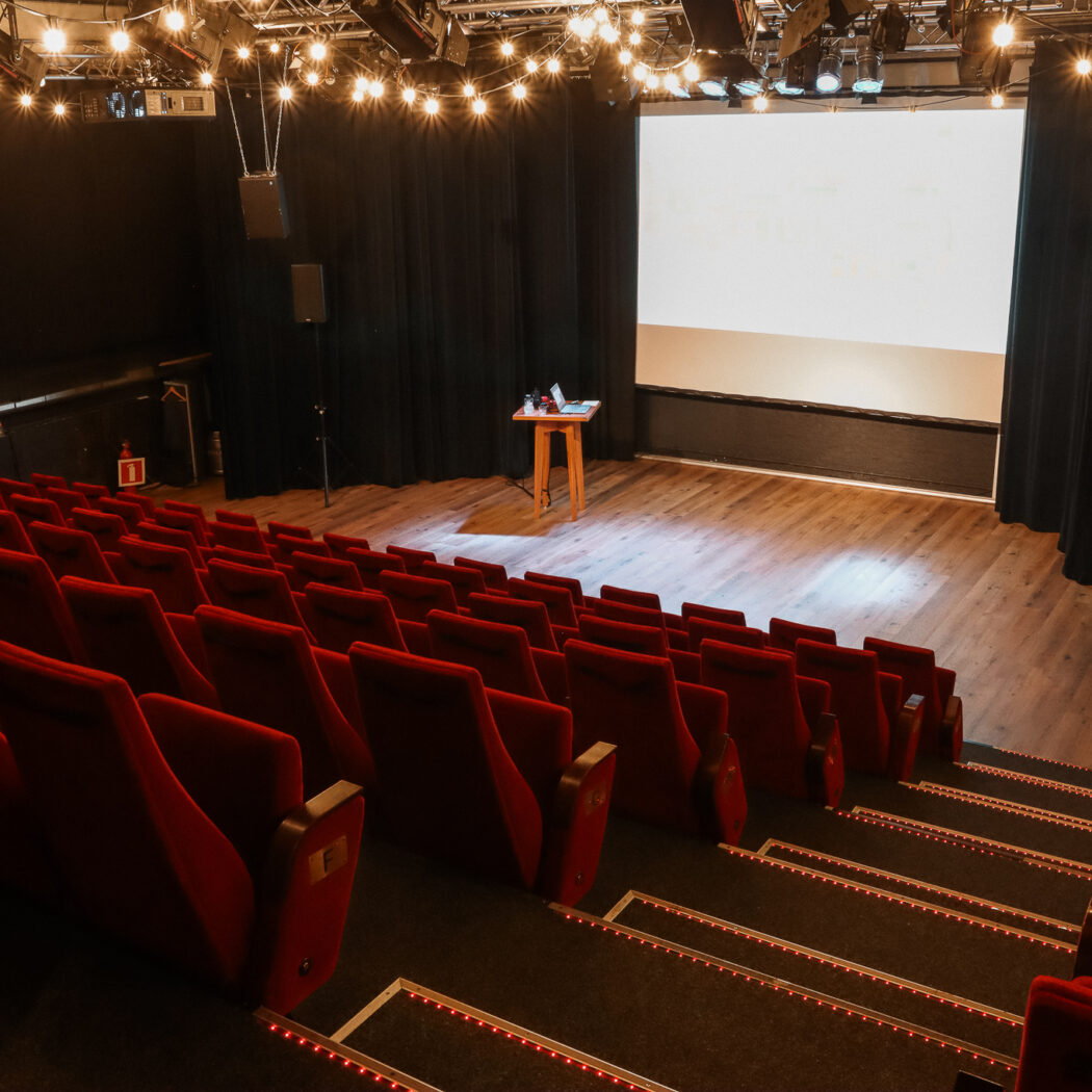 Lege theaterzaal met rijen rode stoelen, houten podium, wit projectiescherm en sfeervolle podiumverlichting.