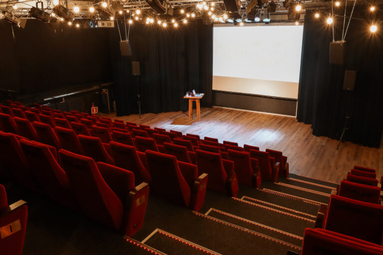 Lege theaterzaal met rijen rode stoelen, houten podium, wit projectiescherm en sfeervolle podiumverlichting.