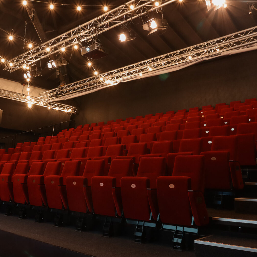 Rode stoelenrijen in een lege theaterzaal met podiumverlichting aan metalen spanten aan het plafond.