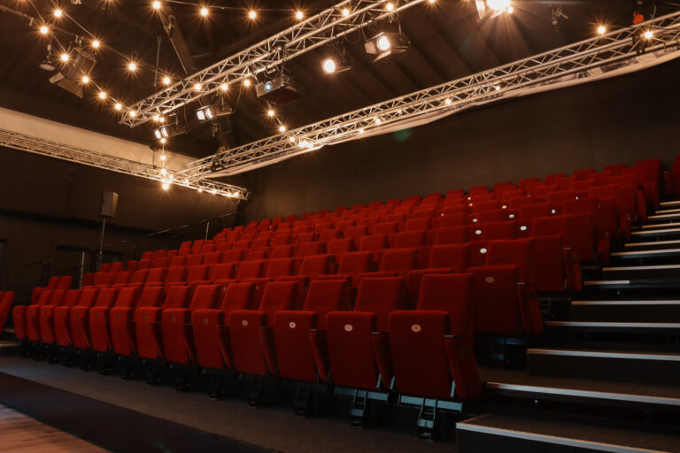 Rode stoelenrijen in een lege theaterzaal met podiumverlichting aan metalen spanten aan het plafond.