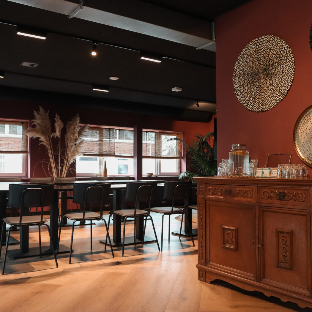 Stijlvolle bijeenkomstlocatie met lange zwarte tafels, roodbruine muren en een sierlijk houten dressoir met water en glazen.