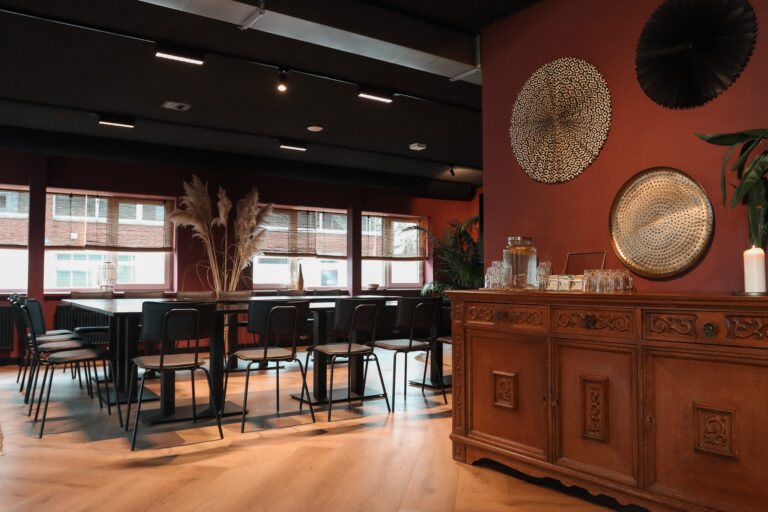 Stijlvolle bijeenkomstlocatie met lange zwarte tafels, roodbruine muren en een sierlijk houten dressoir met water en glazen.