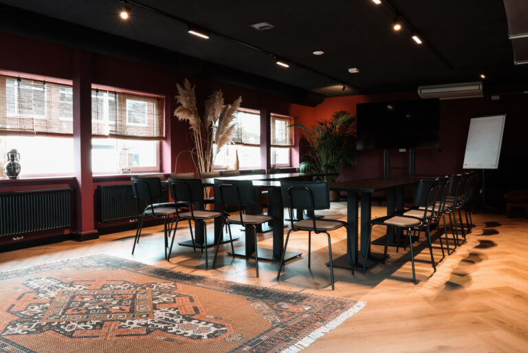 Sfeervolle vergaderruimte met donkerrode muren, houten vloer, zwarte tafels, stoelen, een groot scherm en oranje tapijt.