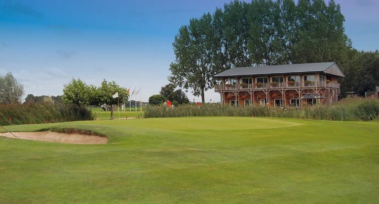 Groene golfbaan met zandbunker en houten clubhuis met balkons en bloembakken onder blauwe lucht.