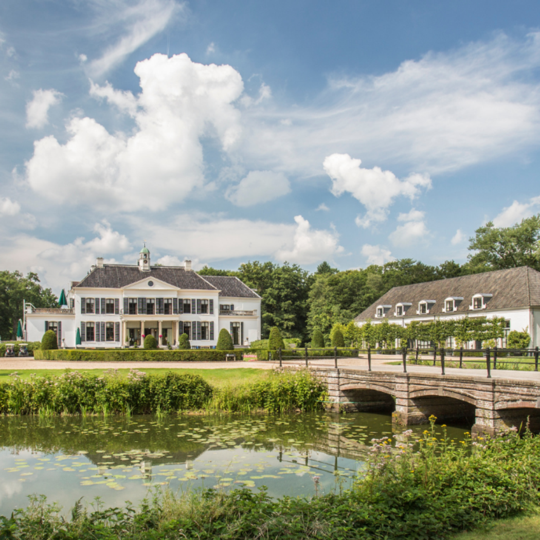 Wit landhuis met donkere luiken, stenen brug over vijver, omringd door groen en een klassiek beeld.