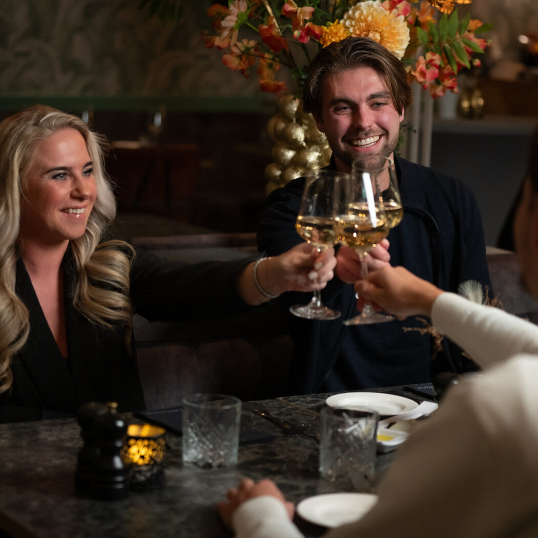 Drie mensen heffen lachend witte wijnglazen aan een tafel in een restaurant.