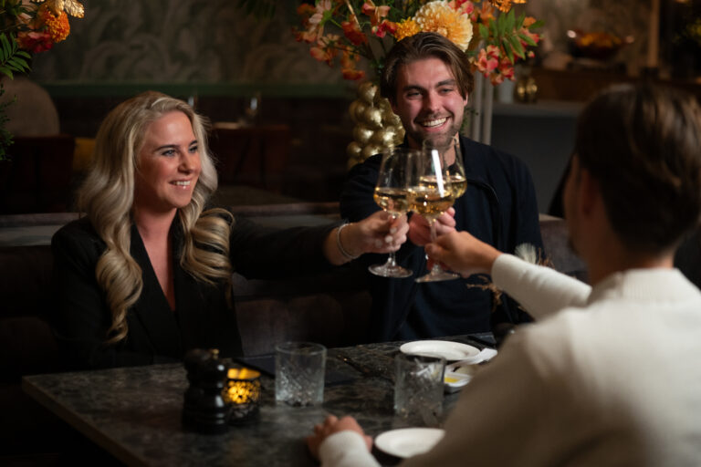 Drie lachende mensen proosten met glazen witte wijn in een sfeervol restaurant.