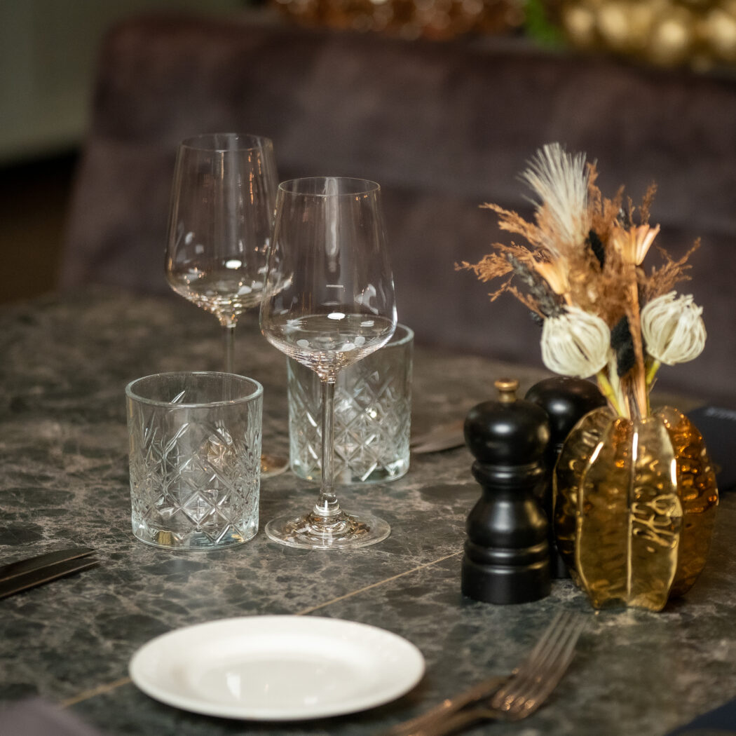 Gedekte donkere marmeren tafel met wijnglazen, gedecoreerde tumblers, gouden vaas met droogbloemen en bestek.