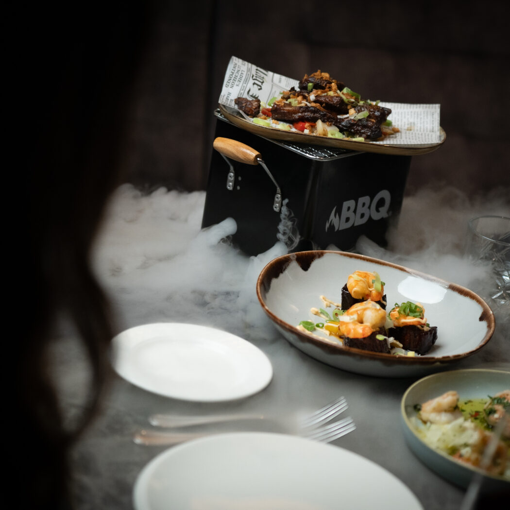 Tafel vol met gerechten: ribbetjes op een mini BBQ en garnalen op donkere hapjes, omringd door dramatische rook.