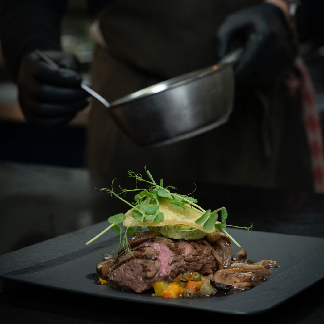 Een chef met zwarte handschoenen giet saus over medium-rare biefstuk met paddenstoelen en microgreens op een zwarte plaat.