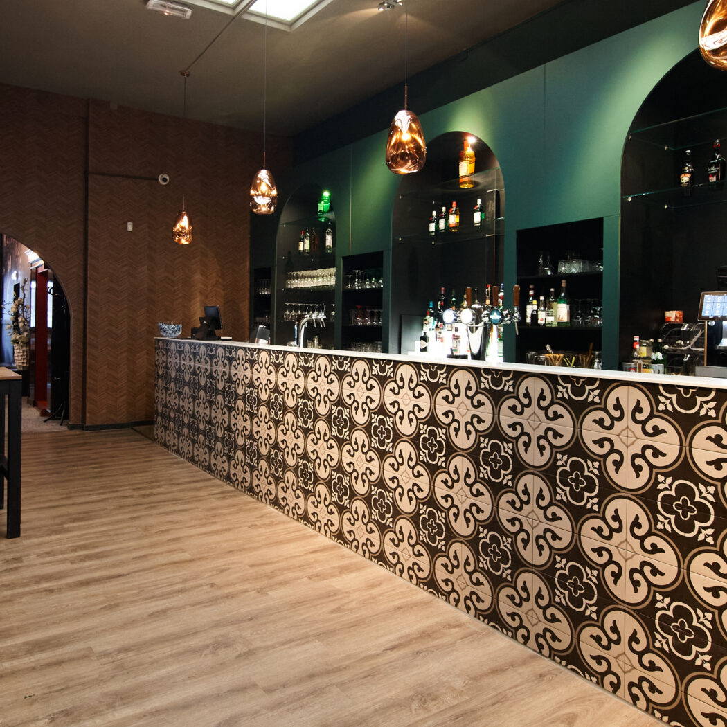 Moderne bar met patroontegels, donkergroene muren en koperkleurige lampen, grenzend aan een eethoek.