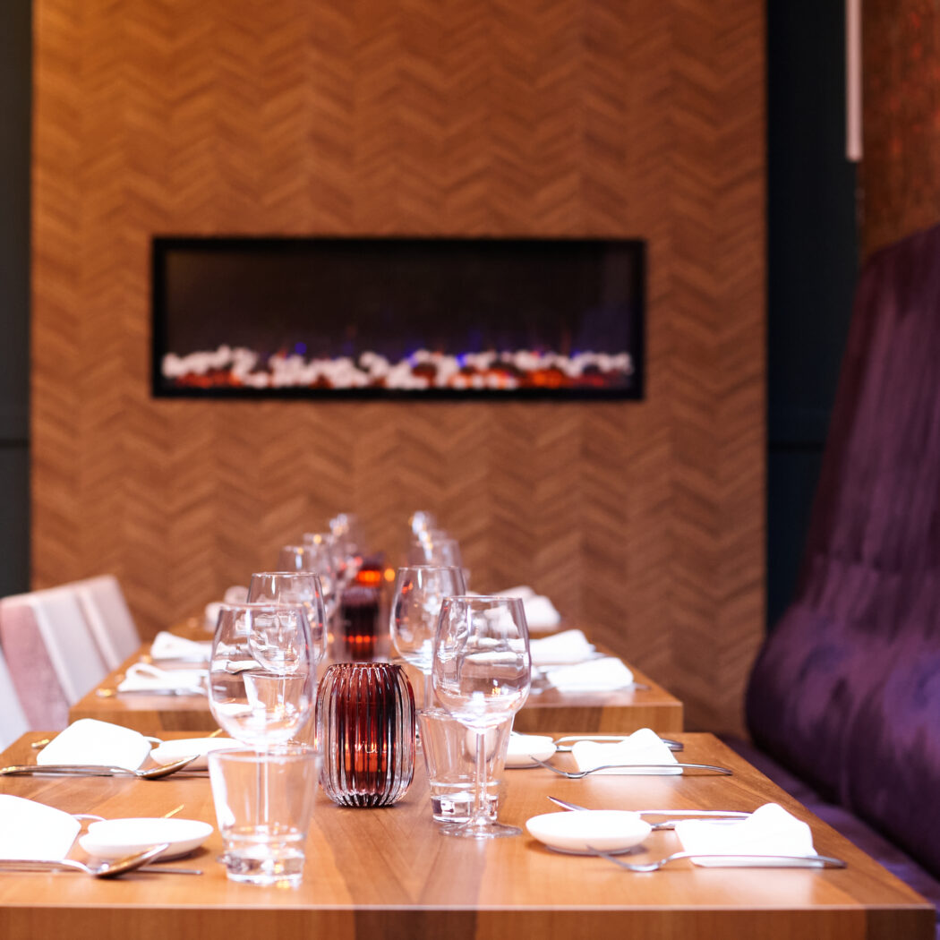 Sfeervolle restauranttafel gedekt met glazen en servies, naast een paarse bank en een moderne sfeerhaard.