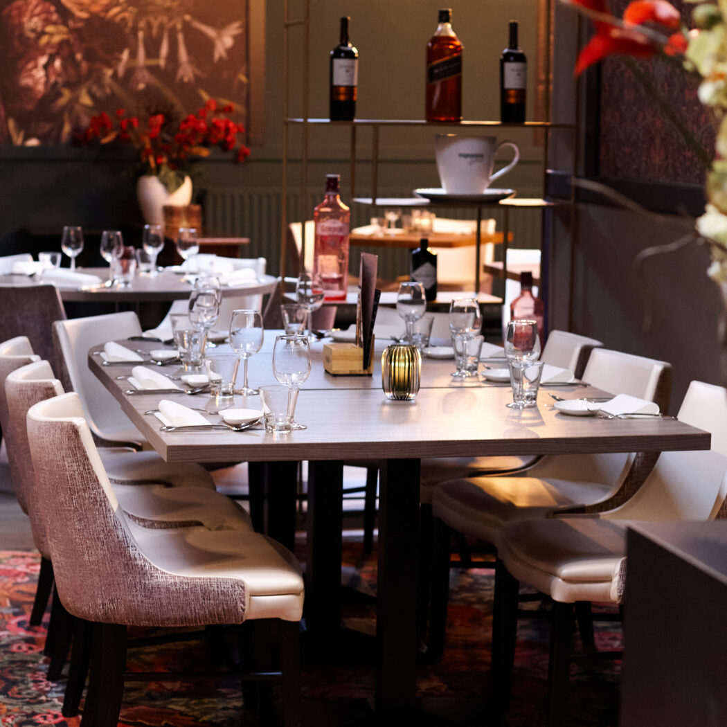 Gedekte rechthoekige restauranttafel met warme sfeerverlichting, bloementapijt en decoratieve flessen op rek.